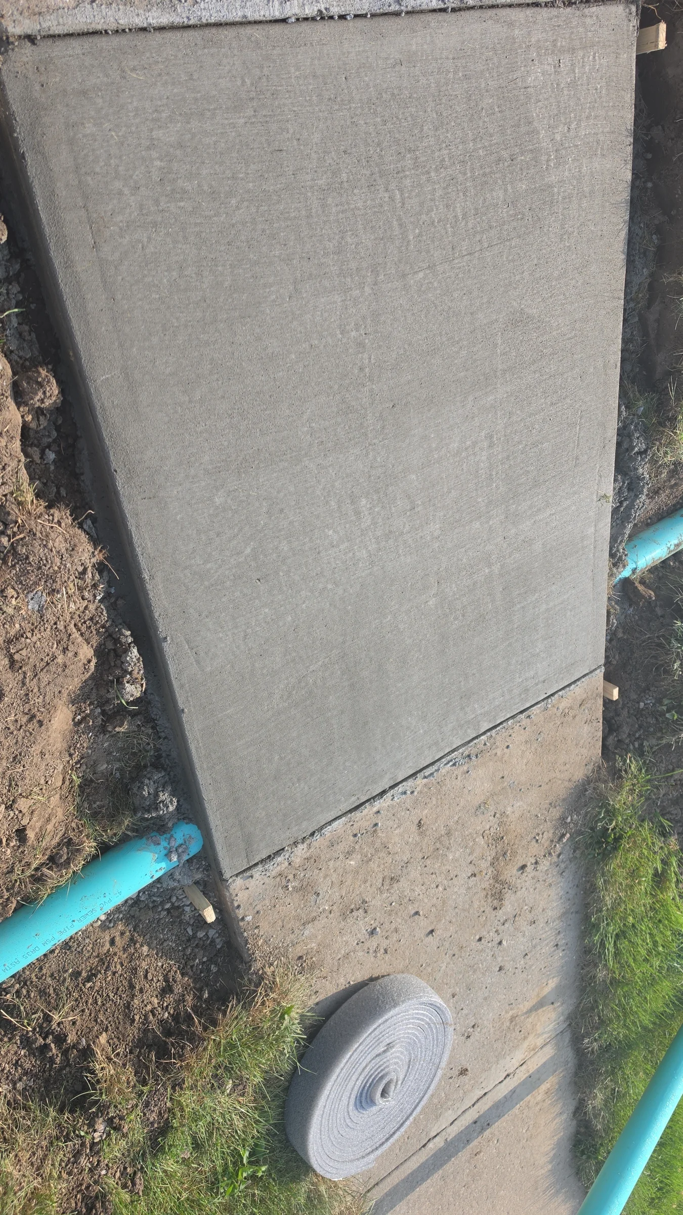 Newly poured concrete walkway with exposed soil, grass, and blue utility pipes around the work area